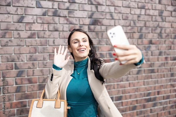 Obraz A joyful woman waves at her selfie while posing confidently outdoors, radiating happiness and energy in a chic outfit.