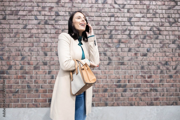 Obraz This image features an elegant woman joyfully conversing on her phone, showcasing her fashionable attire and cheerful demeanor.