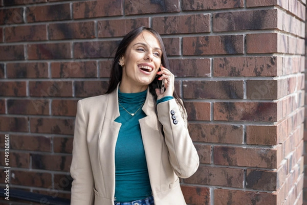 Obraz A lively woman smiles as she talks on her phone against a brick wall, exuding confidence and engagement with her surroundings.