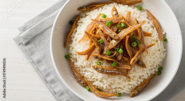 Fototapeta Bowl of rice topped with fried anchovies and green onion garnish, displayed on a gray cloth and white wood background, studio shot, overhead view.