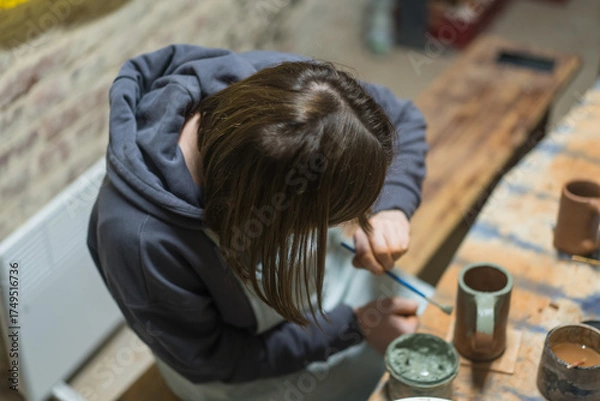 Fototapeta Girl paints her own handmade clay mug. Pottery teaching lesson. Potter makes dishes from clay, ceramics. Top view.