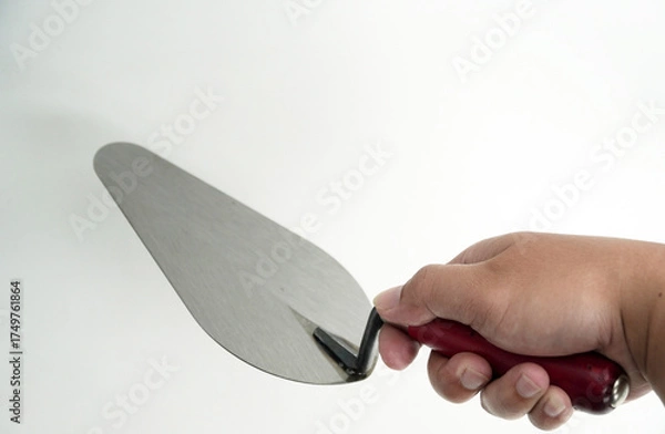 Fototapeta A person's hand firmly grips a professional bricklaying trowel with a red handle, preparing for work. The tool and hand are shown on a clean white background.
