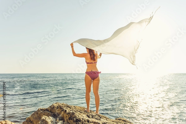 Obraz A girl heaved up hands and the sarong pareo develops in the wind against the blue sky.. On a background the sea. The concept of summer vacations and travel.
