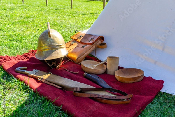 Obraz Set of historical Roman soldier items by the tent — helmet, belt, sword, horn, leather bag and wooden bowls on a red blanket.