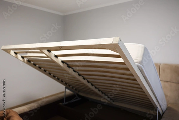Fototapeta  Lift-up bed frame with open storage mechanism in a modern bedroom. Wooden slats and metal supports visible. Practical furniture design and smart home interior concept. High quality photo