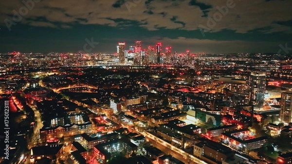 Fototapeta England, London: Canary Wharf and surrounding residential areas buildings skyscrapers glow with colorful lights at night, creating a stunning cityscape view from above, captured by a drone