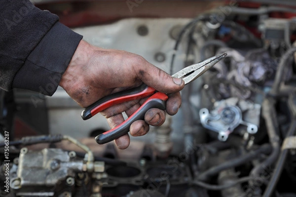 Fototapeta Dirty mechanic hand  hold long nose pliers in hand with car engine in background
