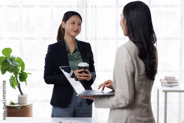 Fototapeta Two business women professionals are working together, using a laptop and discussing work related matters in a modern office setting
