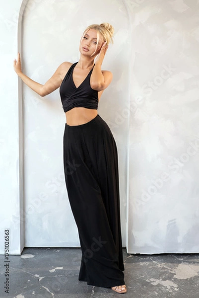 Fototapeta Confident blonde woman posing in a minimalist studio, dressed in a black halter top and wide-leg pants. Fashion-forward, urban look with subtle sensuality. Ideal for fashion