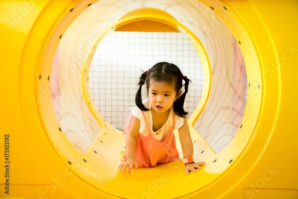 Fototapeta A little smile girl is crawling out of yellow tunnel at playground
