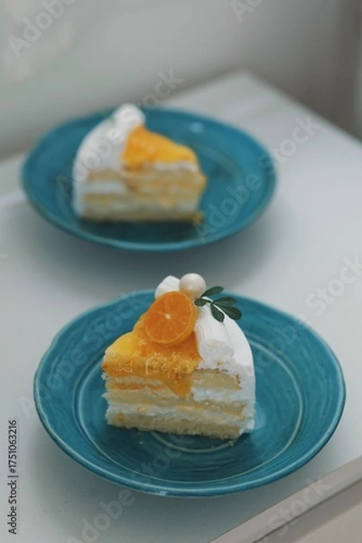 Obraz Close-up of a delicious homemade orange cake topped with whipped cream, orange glaze, and fresh tangerine slice on white background with a cake slice on a blue plate .