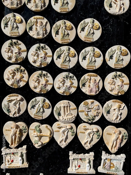 Obraz Greek souvenir magnets depicting ancient mythology — gods, warriors, and temples. Relief-style designs of Leonidas, Zeus, and Olympia, crafted in beige and gold tones on a black display background.