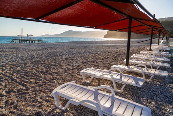 Obraz White plastic lounge chairs are lined up under shade on the beach. Beach umbrellas and sun loungers line the green-painted wooden walkways and wooden pier.
