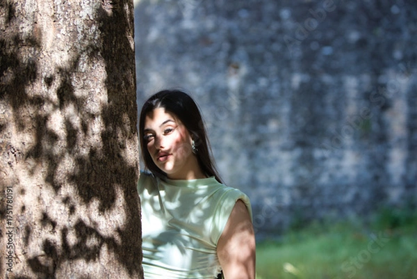 Obraz A beautiful Latin teenager with long dark hair rests her hands and face on a tree trunk in the sunlight. The woman is sad and depressed and looks in different directions.
