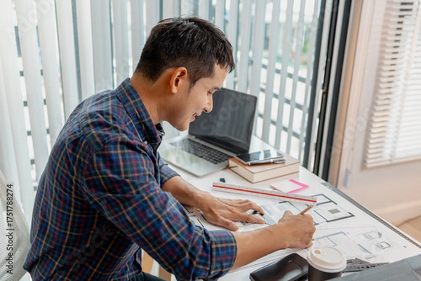 Fototapeta A young male architect is working with blueprints at his desk, using rulers and drawing tools to refine details. He appears focused and engaged, balancing creativity with precision.
