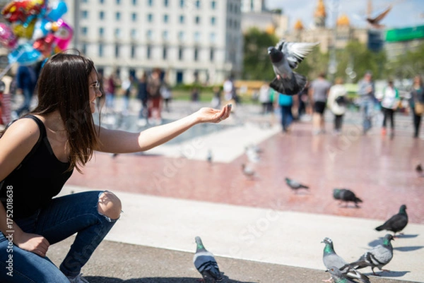 Obraz Joyful young woman feeding pigeons in a sunny city square, enjoying a peaceful moment surrounded by urban life and historical architecture on a vibrant afternoon adventure