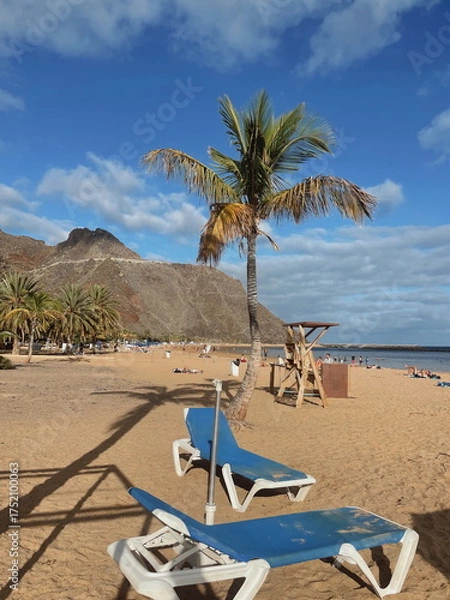 Fototapeta Sun loungers under a palm tree on a tropical beach with golden sand. Perfect vacation scene, peaceful and relaxing coastal atmosphere.