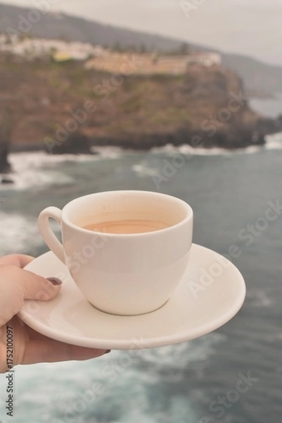 Fototapeta Morning coffee cup with ocean and rocky cliffs in the background. Peaceful coastal view, perfect for travel, relaxation, and lifestyle themes.