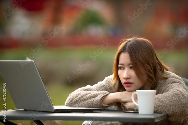 Fototapeta A young Asian woman, dressed in a cozy fleece, appears deeply contemplative or perhaps a bit bored, leaning on her arms at an outdoor table with a laptop and a white coffee mug. The soft, blurred back