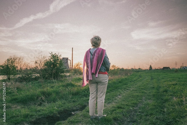 Fototapeta Middle age woman stand alone on coutryside on the path on her back practising mindfullness, relaxing, in the background chimneys, vintage style photo with grain