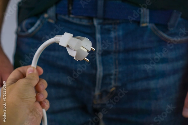 Obraz hand holding a white european power plug with a man in blue jeans and a dark shirt in the background concept of male sexual health, energy and power, erectile dysfunction