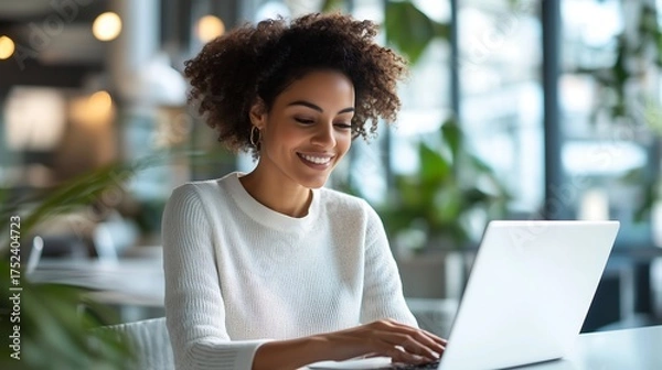 Fototapeta Smiling woman working on laptop in bright modern office, achieving success with ease