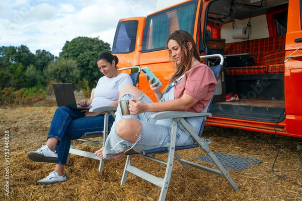 Fototapeta Women relaxing on chairs working with laptop and phone outside camper van.