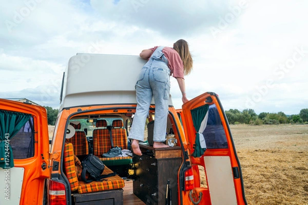 Fototapeta Woman climbing barefoot onto the roof of an orange camper van to clean or check something