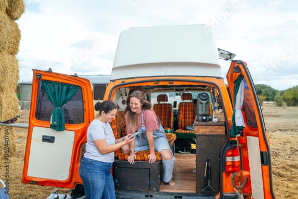 Fototapeta Two women looking at phone beside camper van and hay bales.