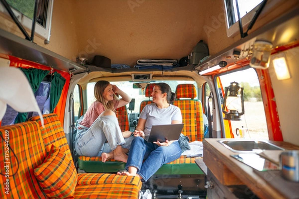 Fototapeta : Two women relaxing inside a camper van, sitting on orange plaid seats, talking and working on a laptop.