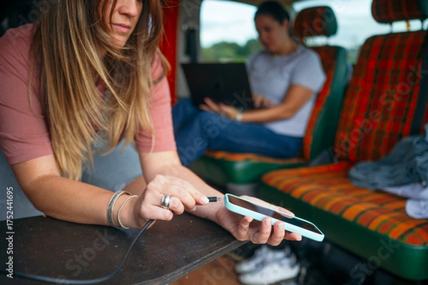 Fototapeta Woman laughing while plugging a charging cable into her smartphone inside a camper van.