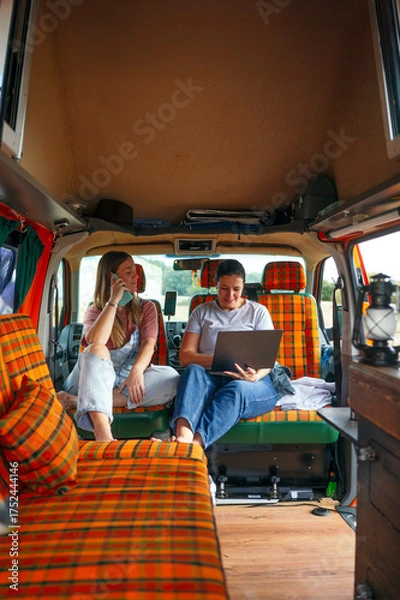 Fototapeta : Two women relaxing inside a camper van, sitting on orange plaid seats, talking and working on a laptop.