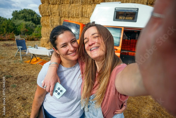 Fototapeta Two women smiling taking a selfie together near orange camper van.