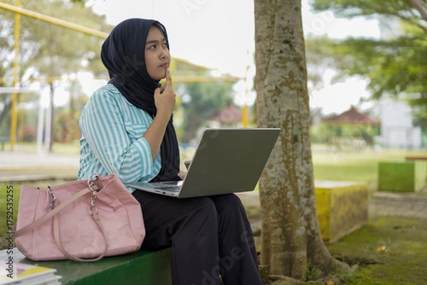 Fototapeta Young Muslim woman wearing a hijab diligently studying outdoors on a park bench with her laptop and books, embracing a modern lifestyle of remote learning and higher education