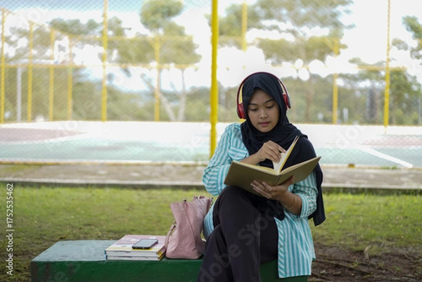 Obraz Serene young woman in a hijab and red headphones, deeply engrossed in reading a book while studying outdoors on a tranquil park bench, fostering knowledge and personal growth