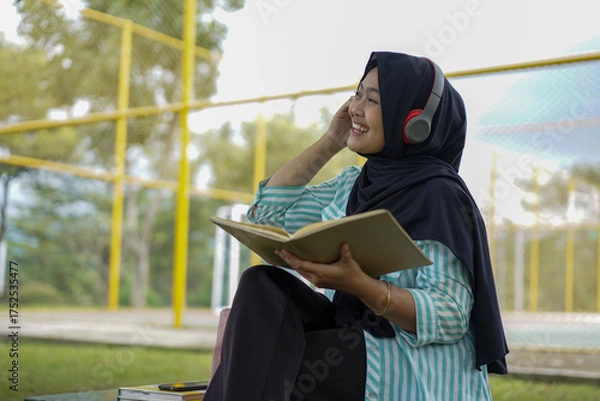 Obraz Serene young woman in a hijab and red headphones, deeply engrossed in reading a book while studying outdoors on a tranquil park bench, fostering knowledge and personal growth