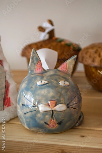 Fototapeta colorful ceramic cat sculpture sits proudly on wooden shelf. its playful features are highlighted by soft colors, surrounded by other handcrafted items, creating charming display. vertical. close up.