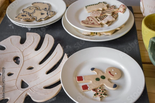 Fototapeta decorative plates hold festive cookies, including gingerbread figures and shapes, arranged on wooden table with tropical leaf design, creating cheerful atmosphere. close up.