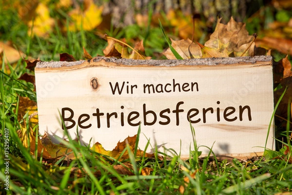 Obraz Betriebsferien Herbst: Holzschild mit hinzugefügtem Text WIR MACHEN BETRIEBSFERIEN auf einer Wiese mit Herbstlaub