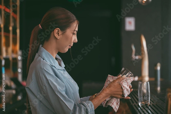Fototapeta Young woman bartender working behind the counter, carefully cleaning a glass with a towel in a modern bar. restaurant industry concept.