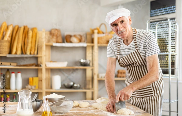 Fototapeta Elderly man baker cutting dough with scraper