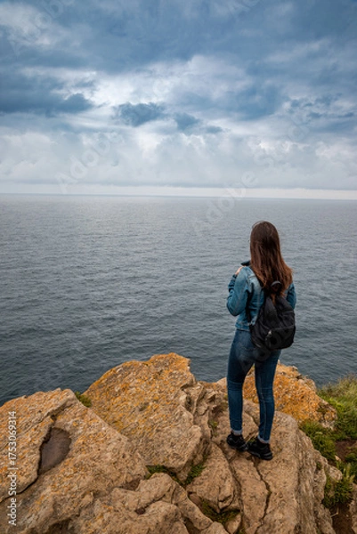 Obraz Contemplative traveler gazes at the vast ocean from dramatic rocky cliff on overcast day, seeking inspiration and adventure on a solo journey along the scenic coastline