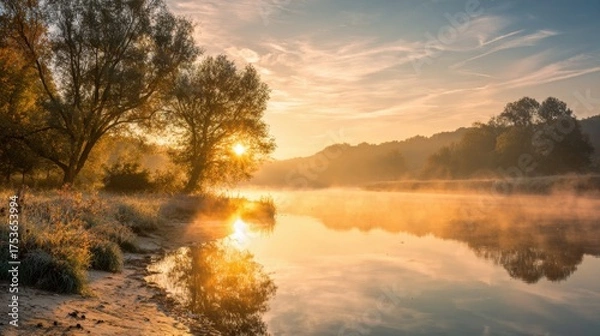 Fototapeta Soft light breaks through trees as the sun rises over a serene river on a foggy morning.