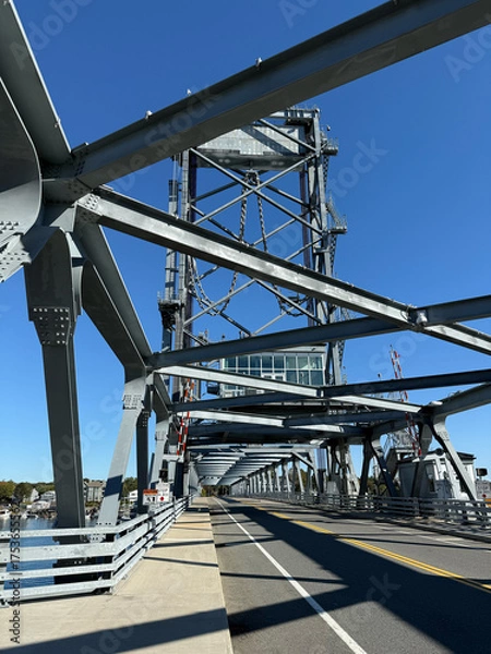 Fototapeta Modern lift bridge, steel truss drawbridge with metal beams, pedestrian walkway, and vehicle lanes extending over calm river