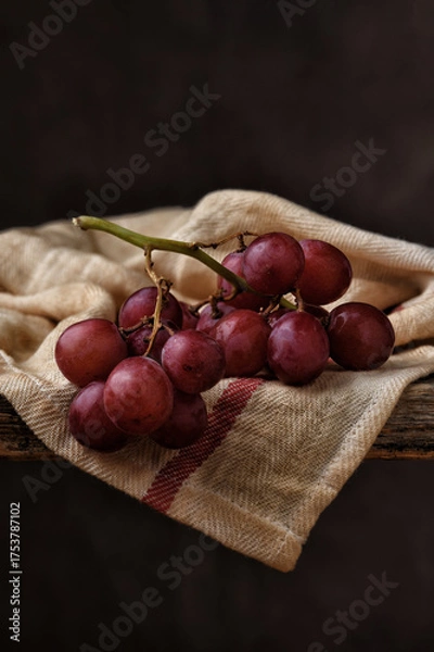 Obraz Red Grape Still Life. Closeup of  a bunch of ripe red grapes on a kitchen towel draped over the edge of a rustic wooden table with a mottled brown background.