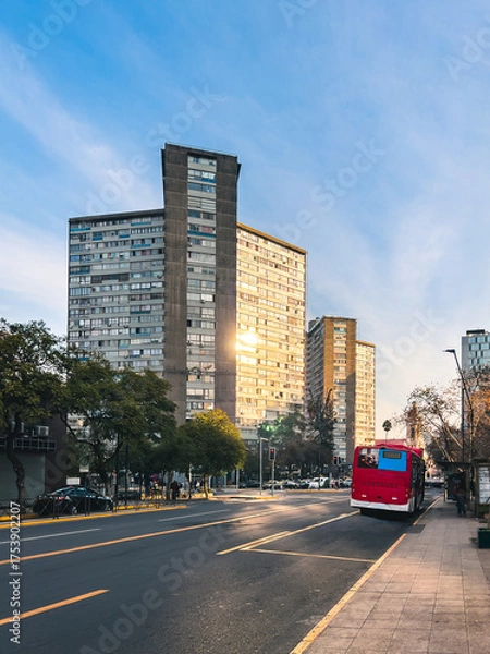 Fototapeta Red city bus driving through urban avenue at sunset, surrounded by tall buildings and trees. Concept of sustainable transport, daily commute, city mobility and modern infrastructure.