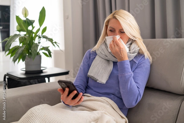 Obraz Middle aged blonde woman feeling unwell, sitting on couch and using her smartphone for work.
