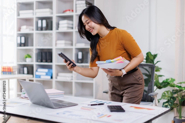 Fototapeta Image of attractive asian woman using mobile phone while sitting at laptop indoor and working with documents