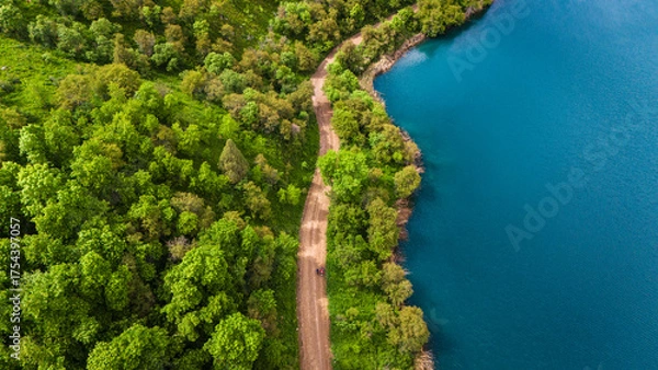 Fototapeta Aerial view of a dirt road running along a turquoise lake surrounded by lush green forest. Scenic natural landscape captured from drone in summer daylight