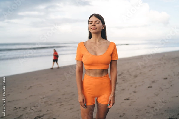 Fototapeta Peaceful brunette in orange sportswear standing still with eyes closed, embracing emotional calmness, digital pause and conscious daily rhythm outdoors.